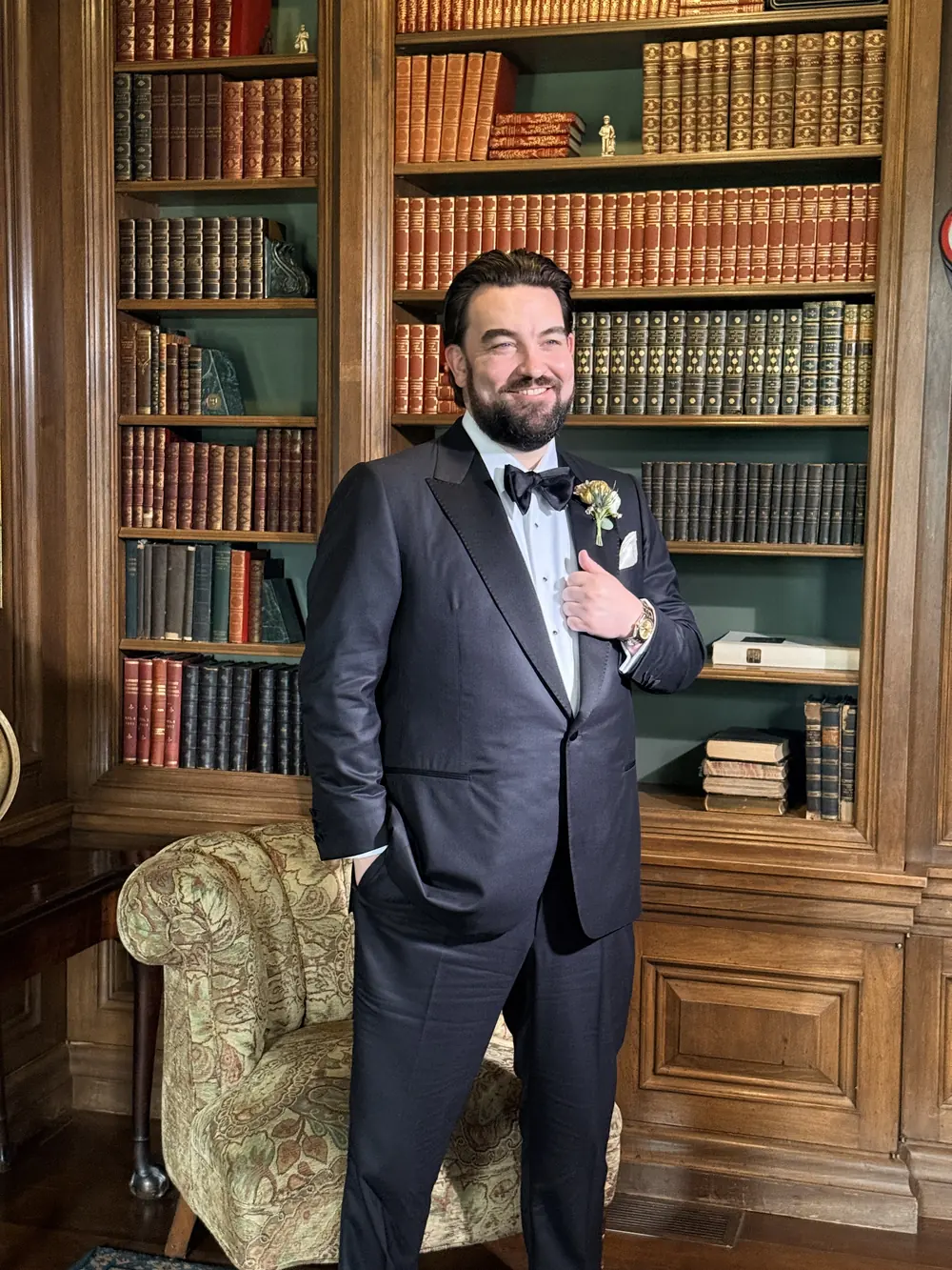 Groom in a navy tuxedo and boutonniere smiling in a wood-paneled library after wedding-morning grooming