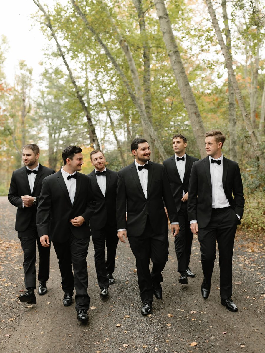 Groom and groomsmen in black tuxedos walking a wooded path on the wedding day