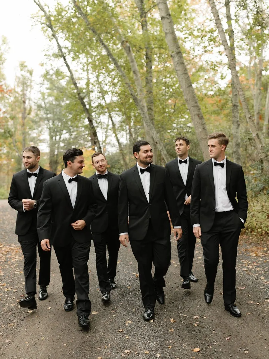 Groom and groomsmen in black tuxedos walking a wooded path on the wedding day