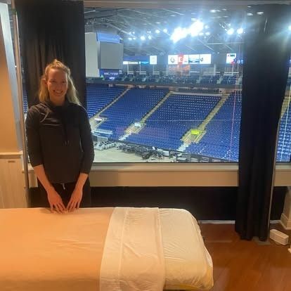 Massage therapist beside a treatment table in an arena suite overlooking the seating bowl