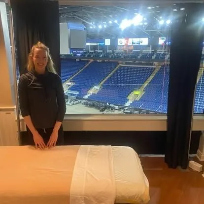 Massage therapist beside a treatment table in an arena suite overlooking the seating bowl